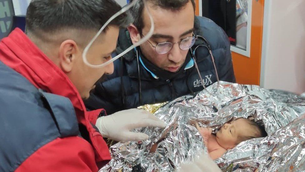 A baby wrapped in a thermal blanket is attended by two men after being rescued from a ruined building
